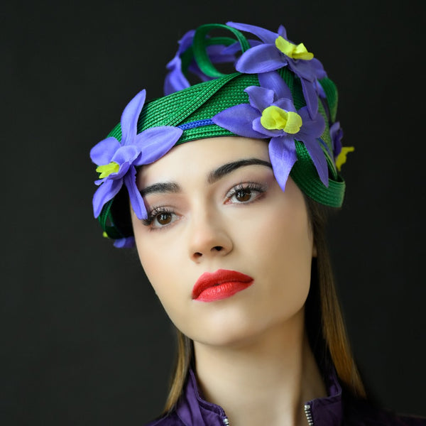 A woman wearing a green dress hat with purple and yellow flower embellishments.