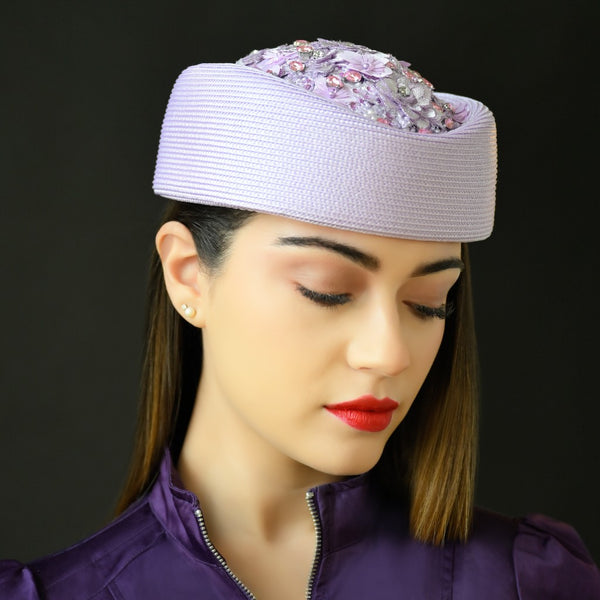 A woman wearing a lavender pillbox hat with lace detailing and embellished with beads and precious stones.