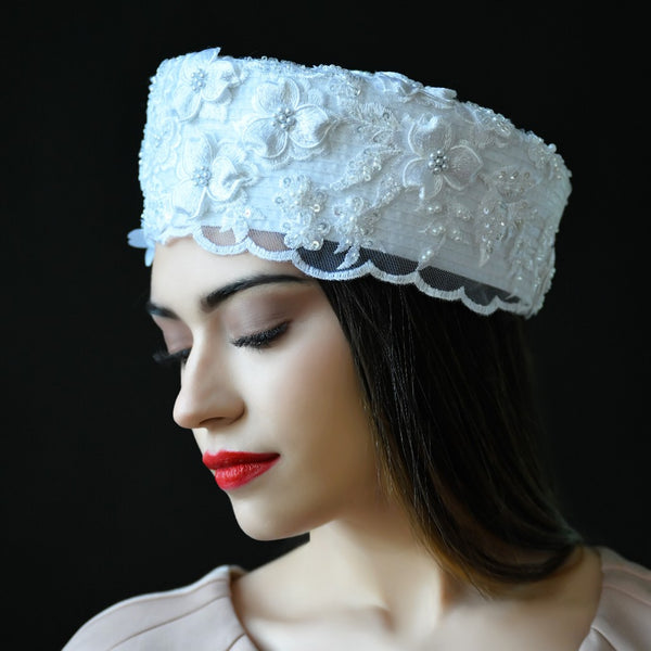 A woman wearing a white lace covered dress hat with beaded accents.