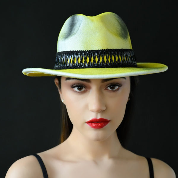 A woman's fedora dress hat with a yellow, black, and cream color scheme and a leather trim. The hat is modeled by a person against a black background.